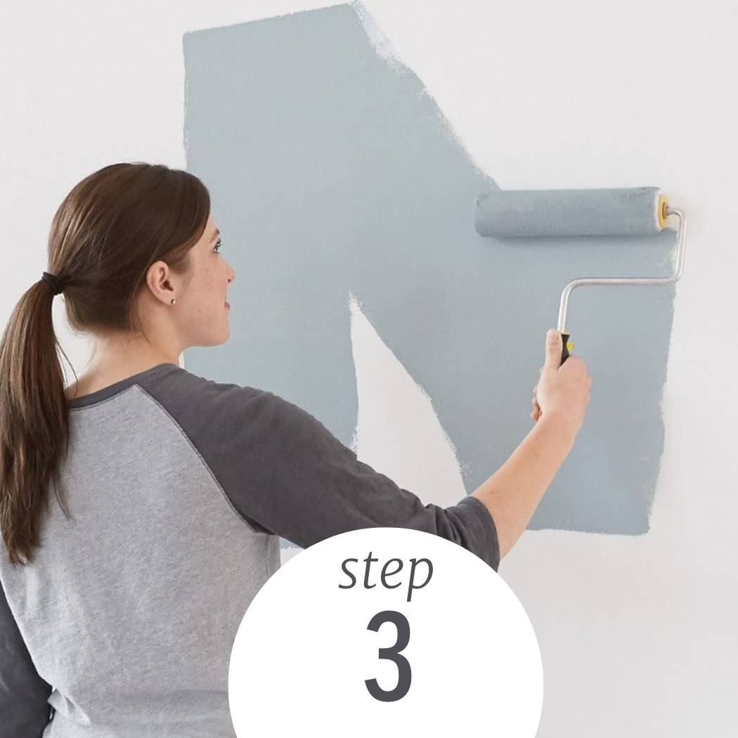 A woman stands at a wall, applying grey paint with a roller brush.