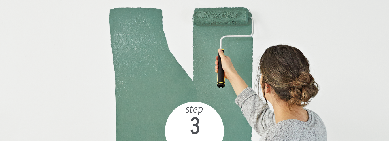 A woman stands at a wall, applying green paint with a roller brush.