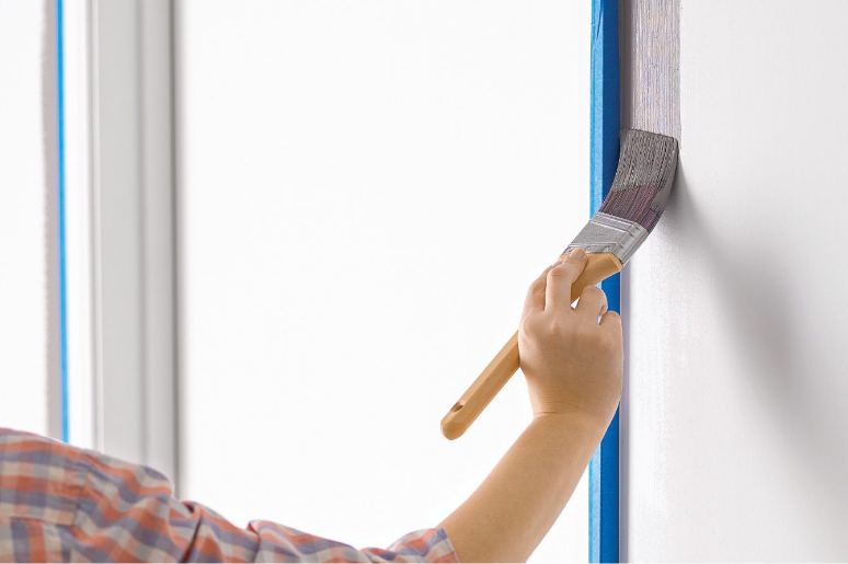 A white wall with blue painter’s tape. A person holds a paintbrush, painting the wall gray. 
