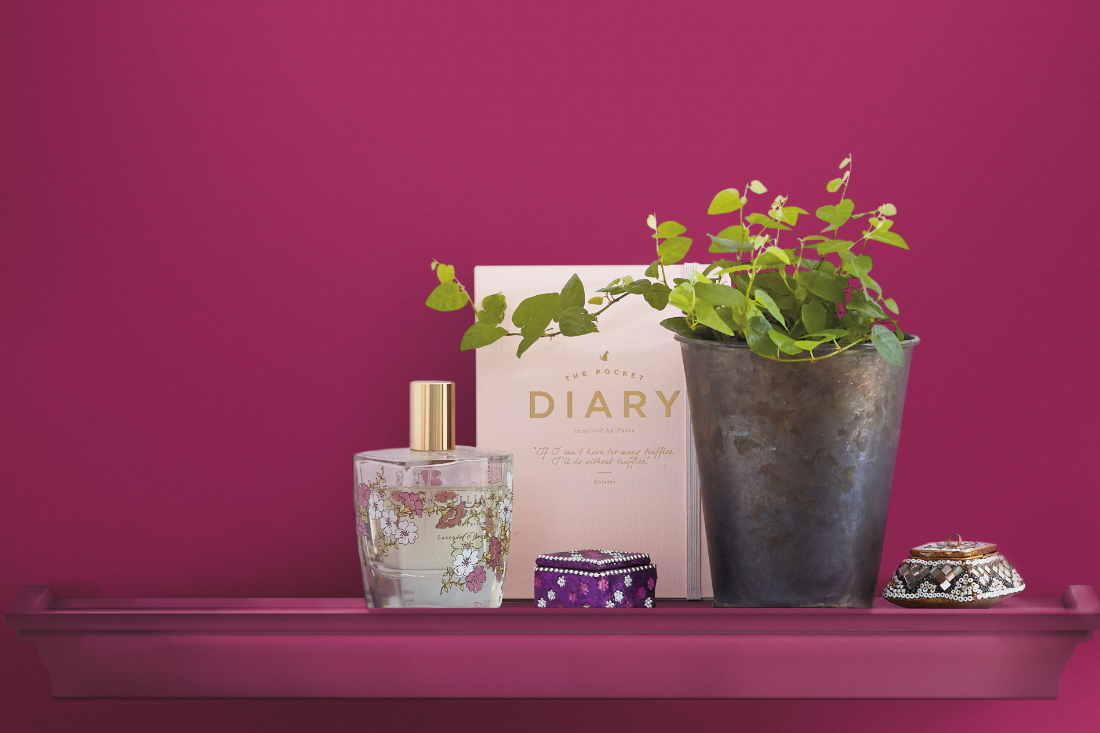 A wall and shelf are painted a deep fuchsia, with a potted plant, a diary, room spray bottle and candle on top of it.