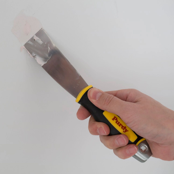 A hand applies putty to a damaged wall using a Purdy putty knife.