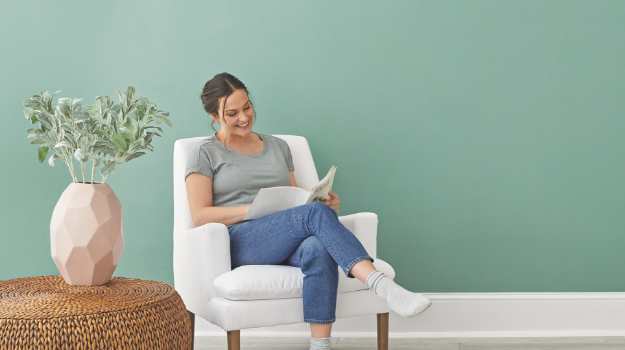 A woman sits in a cozy white accent chair next to a woven side table, reading in her newly painted room.