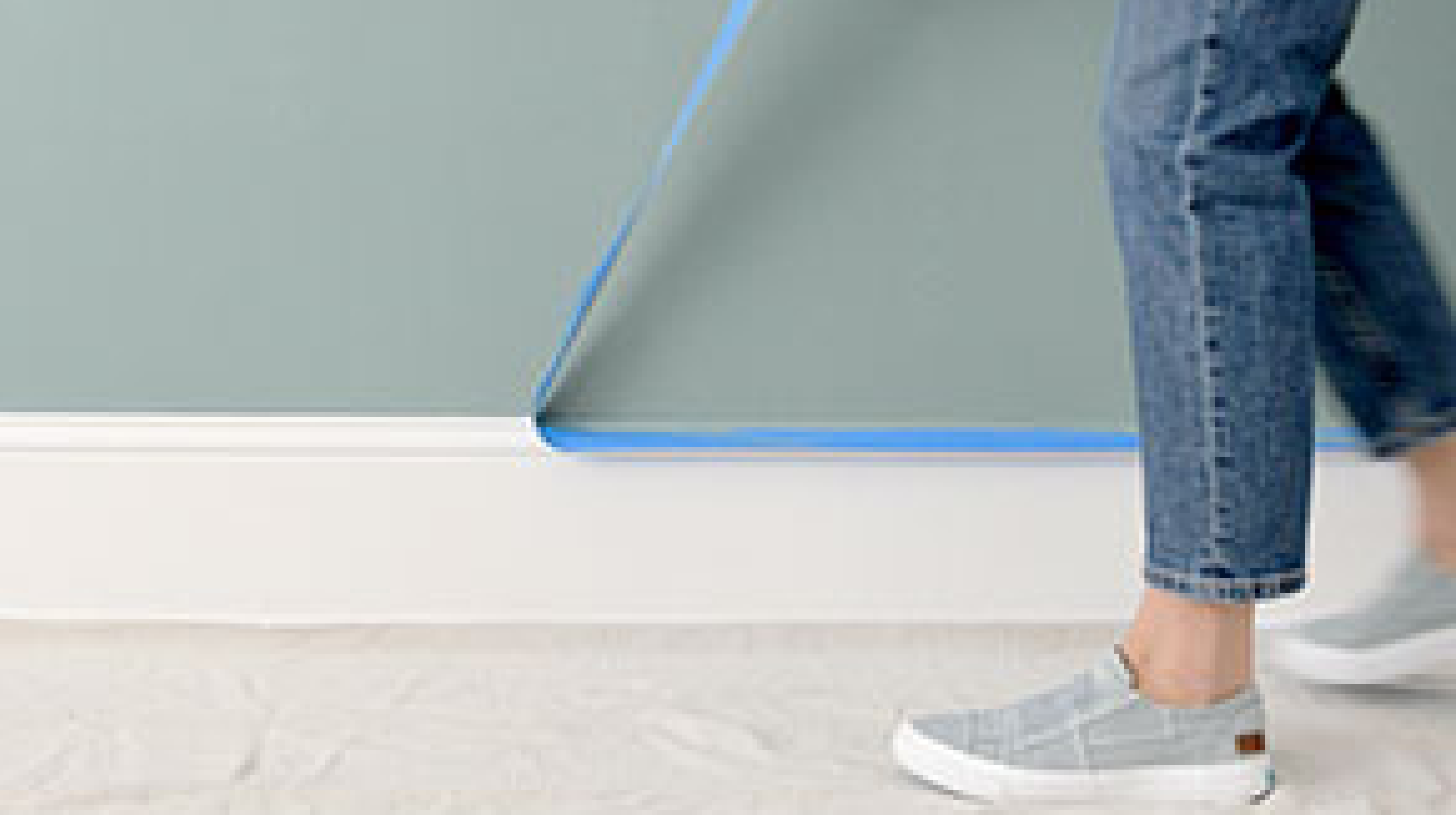 A woman walks along an interior baseboard as she removes painter’s tape from a freshly painted wall.