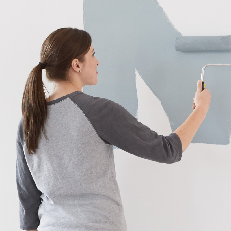 A woman rolls grey paint onto an off-white-colored wall.