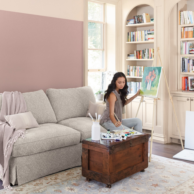A young woman sits on a couch, painting on an easel in a living room, walls painted neutral hennepin stone.