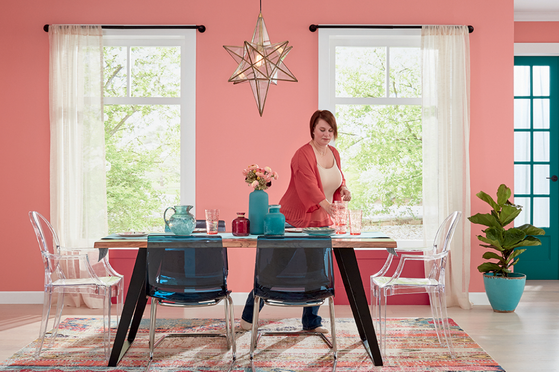 Bright dining room with large windows, trees outside, woman sets dining table. Walls painted Carmela coral. 