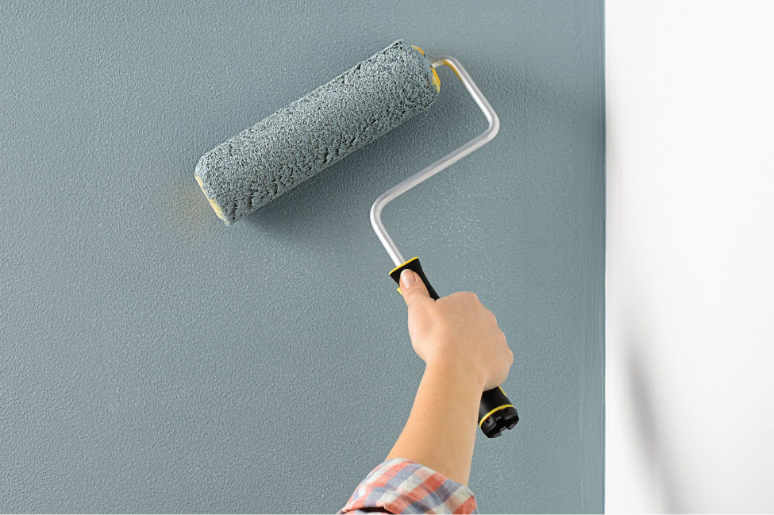 A white wall. A person applies gray paint on the wall with a paint roller.
