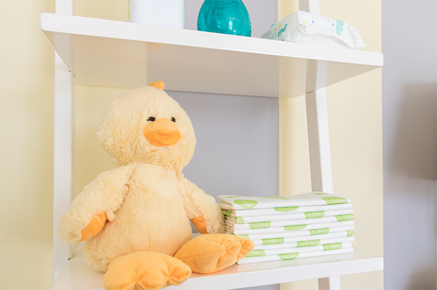 Open bathroom shelves with a stuffed bird and stacked diapers. Wall is vertically striped yellow and grey.